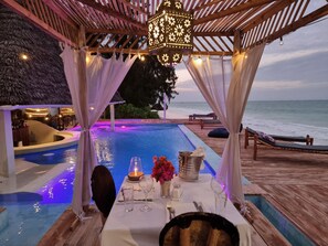 A poolside dining area with a view of the ocean, a wooden deck, and a thatched roof structure. - Blue Palm Zanzibar (Bwejuu)