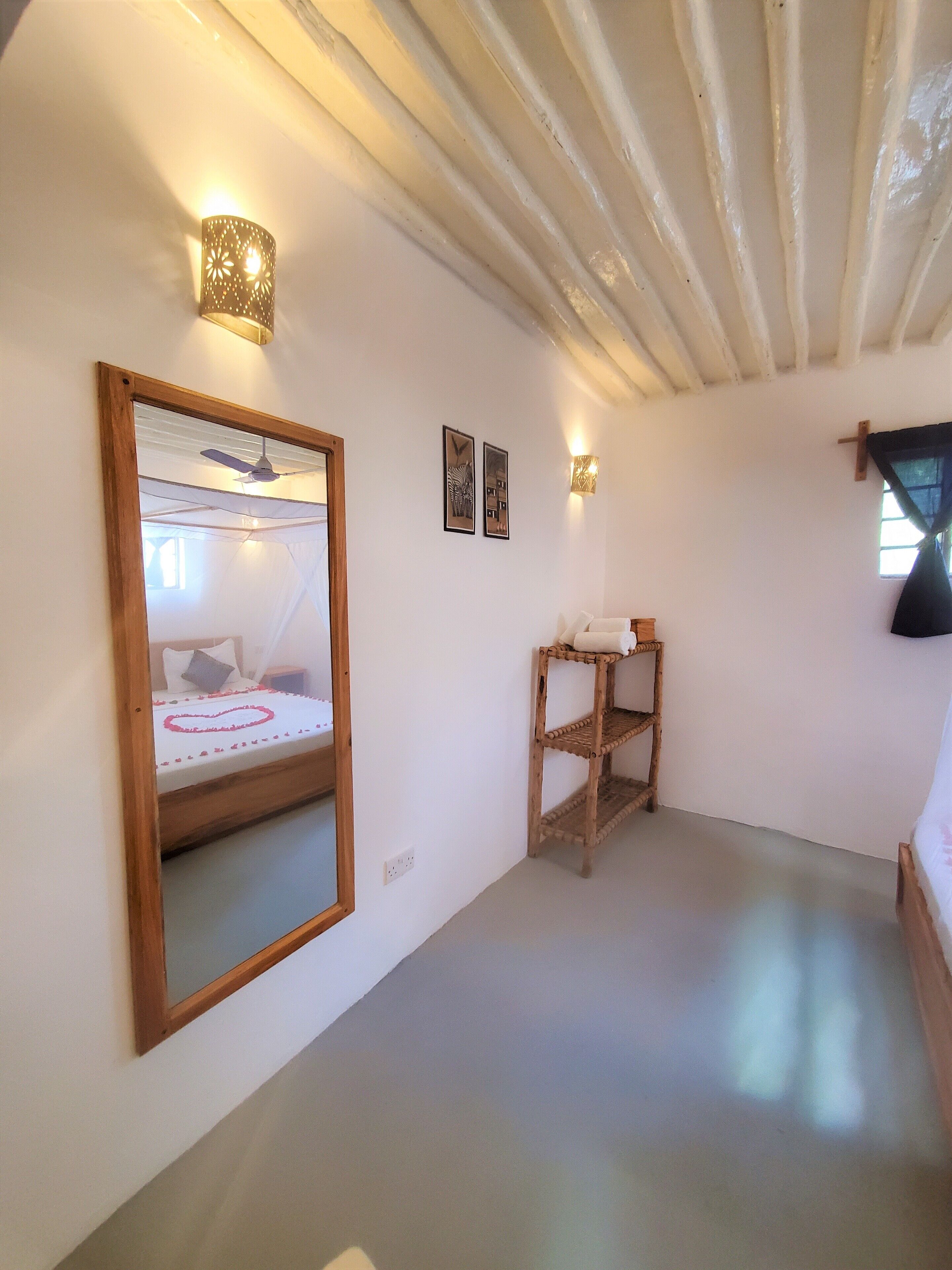Un dormitorio con cama, cabecera de madera, espejo, mesita de noche, dos lámparas de pared y ventilador de techo.