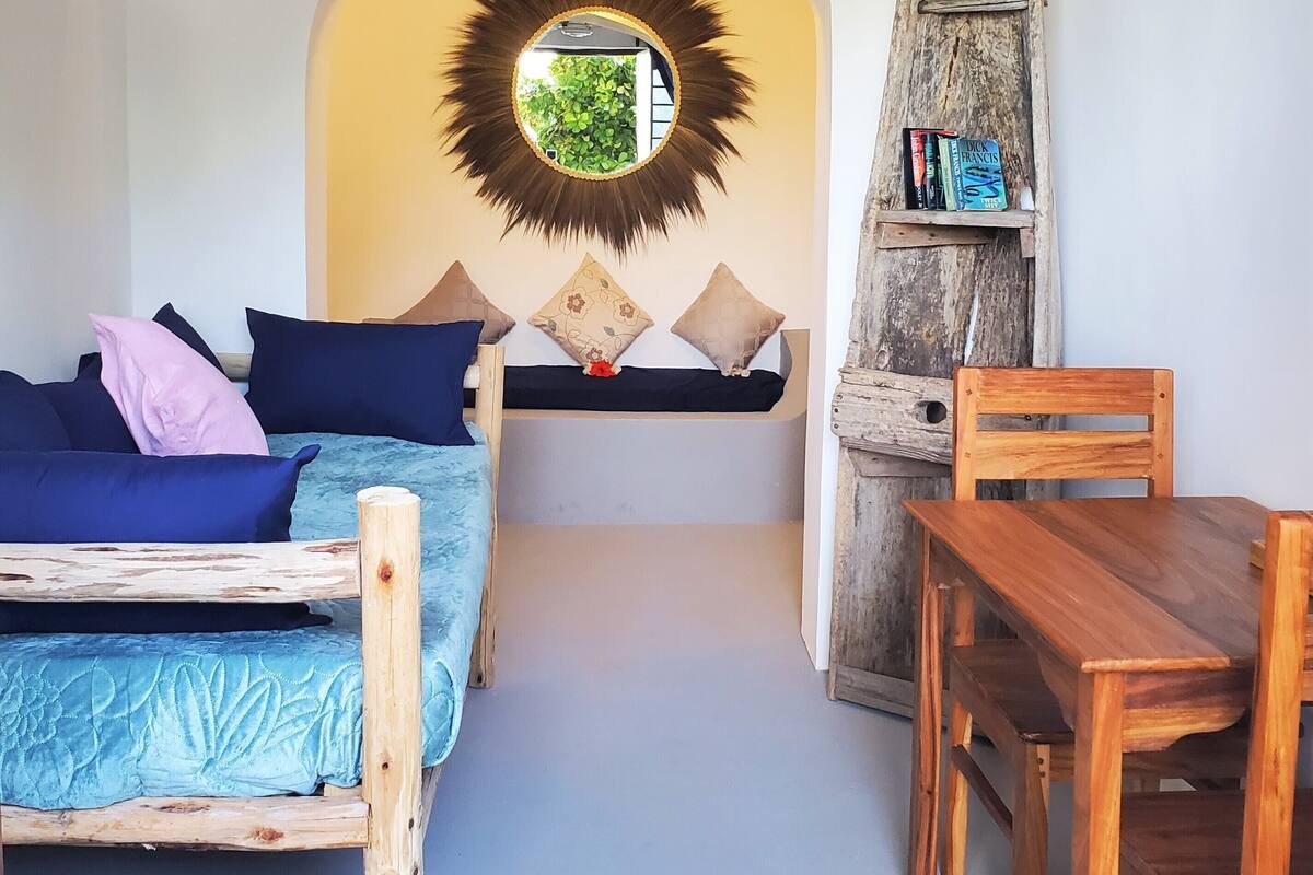 A cozy room with a bed, a wooden table, a chair, a bookshelf, and a ceiling fan.