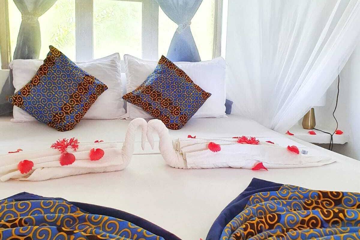 A neatly made bed with patterned pillows and towels, a bedside table with a lamp, and a window with sheer curtains.