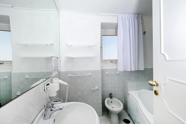 Un baño con bañera, bidé, lavamanos y espejo. Hay barras de apoyo en las paredes y una cortina de ducha.