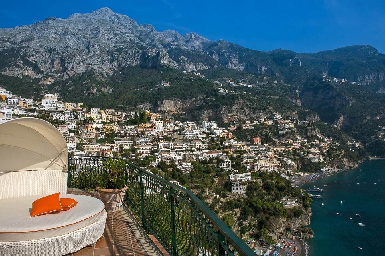 A coastal town with a view of mountains, a beach, and a balcony with a chair and cushions.
