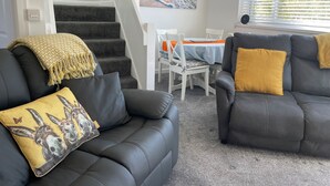 A living room with two black leather sofas, a yellow throw blanket, and a table with chairs. A staircase is visible in the background.