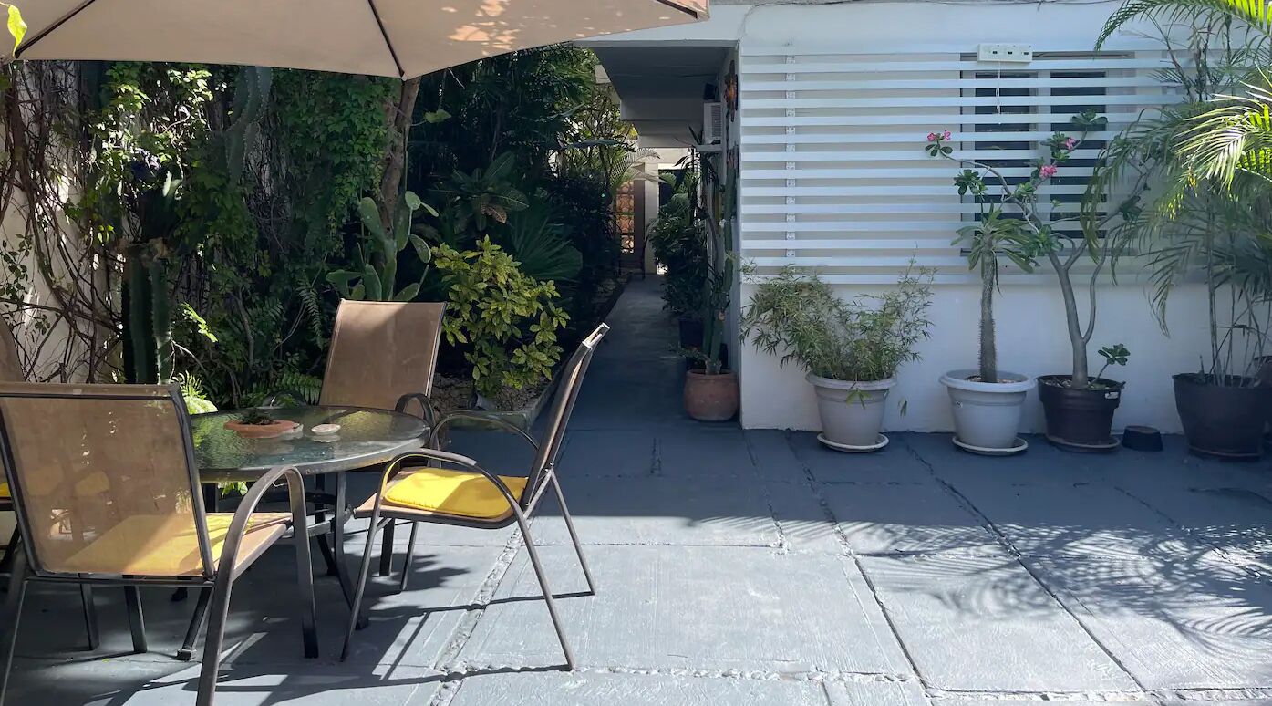 Un patio con mesa y sillas bajo una pérgola, rodeado de plantas en macetas y una pared blanca con persianas horizontales.