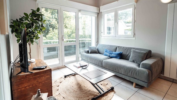 A modern living room with a grey sofa, a coffee table, a TV on a wooden stand, and a large window with a view of greenery. - Cozy apartment by the sea (La Teste-de-Buch)