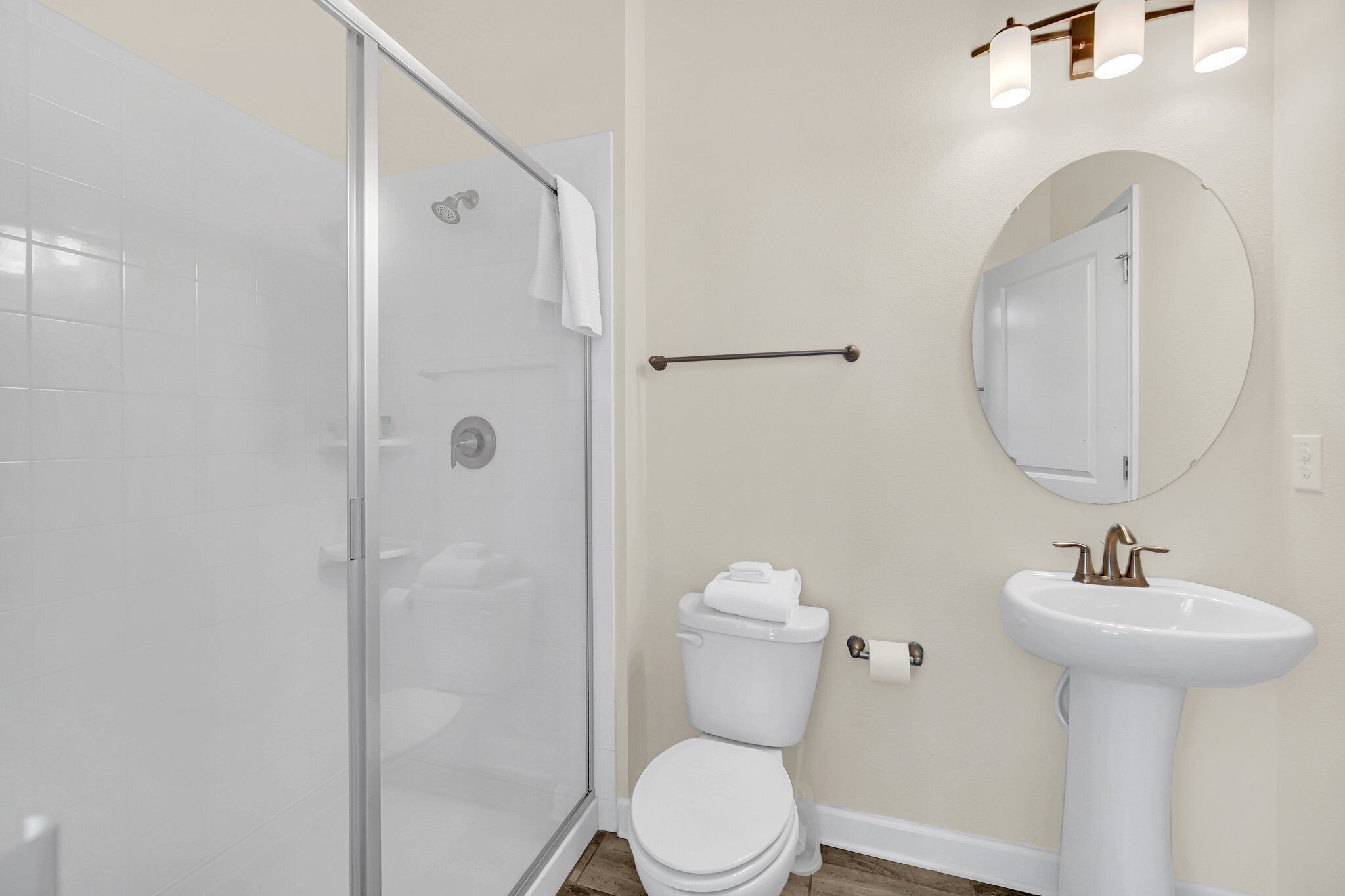 Un baño con cabina de ducha de vidrio, inodoro blanco, lavamanos con grifo dorado y un espejo redondo en la pared.