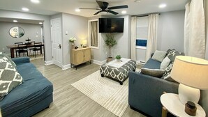 A living room with a ceiling fan, a TV, and a dining area with a table and chairs. - Lakefront on Main  (Elmer)