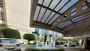 Um edifício moderno com um espelho d'água central, rodeado por uma área ajardinada com arbustos bem cuidados.