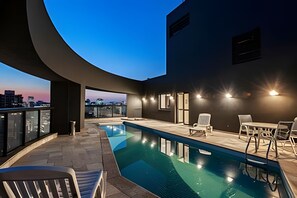 Zona de piscina en la azotea con vista al horizonte urbano al anochecer, mobiliario exterior y diseño arquitectónico moderno.