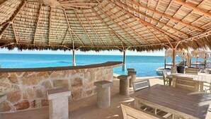 Eine Terrasse am Strand mit einem Reetdach, Steinmauern und Holzmöbeln mit Blick auf den Ozean.