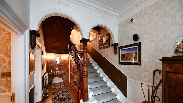 A grand staircase with a wooden handrail and a carpeted floor leading to a room with a fireplace. - The Townhouse (Ayr)