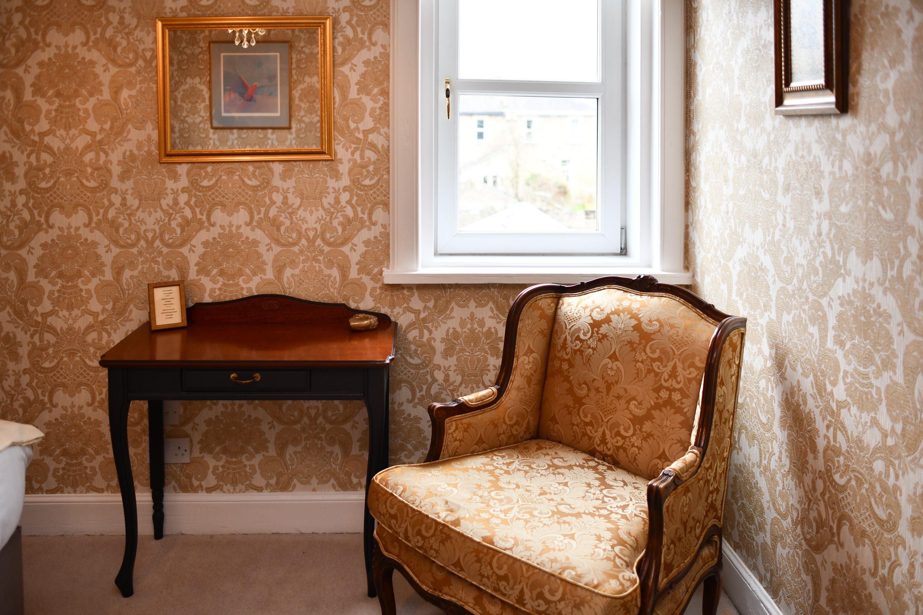 A room with patterned wallpaper, a wooden desk with a drawer, a cushioned armchair, and a window with a view of buildings outside.