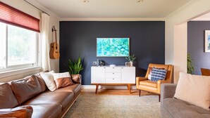 A living room with a brown leather sofa, a white cabinet, a wooden chair, and a large window with red curtains.