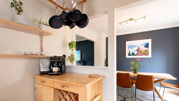 A modern kitchen with a coffee machine, wine rack, and hanging pots. There is a dining area with a table and chairs.
