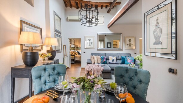 A dining area with a table set for two, a chandelier, and a view into the living room. - Panoramic Penthouse with Terrace, 2 bedrooms 2 bathrooms inside the Walls of Lucca (Lucca)