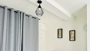 Una habitación con cortinas grises, un banco, dos cuadros enmarcados, un radiador blanco y una lámpara colgante.