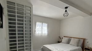 Un dormitorio con cama, una silla, un ventanal con cortinas, una lámpara en el techo y una pared con pequeños ventanales.