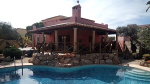 A pool area with a stone border, a wooden deck, and a building with a red facade and columns.