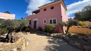 A pink villa with a tiled roof, a small porch, and a gravel driveway.