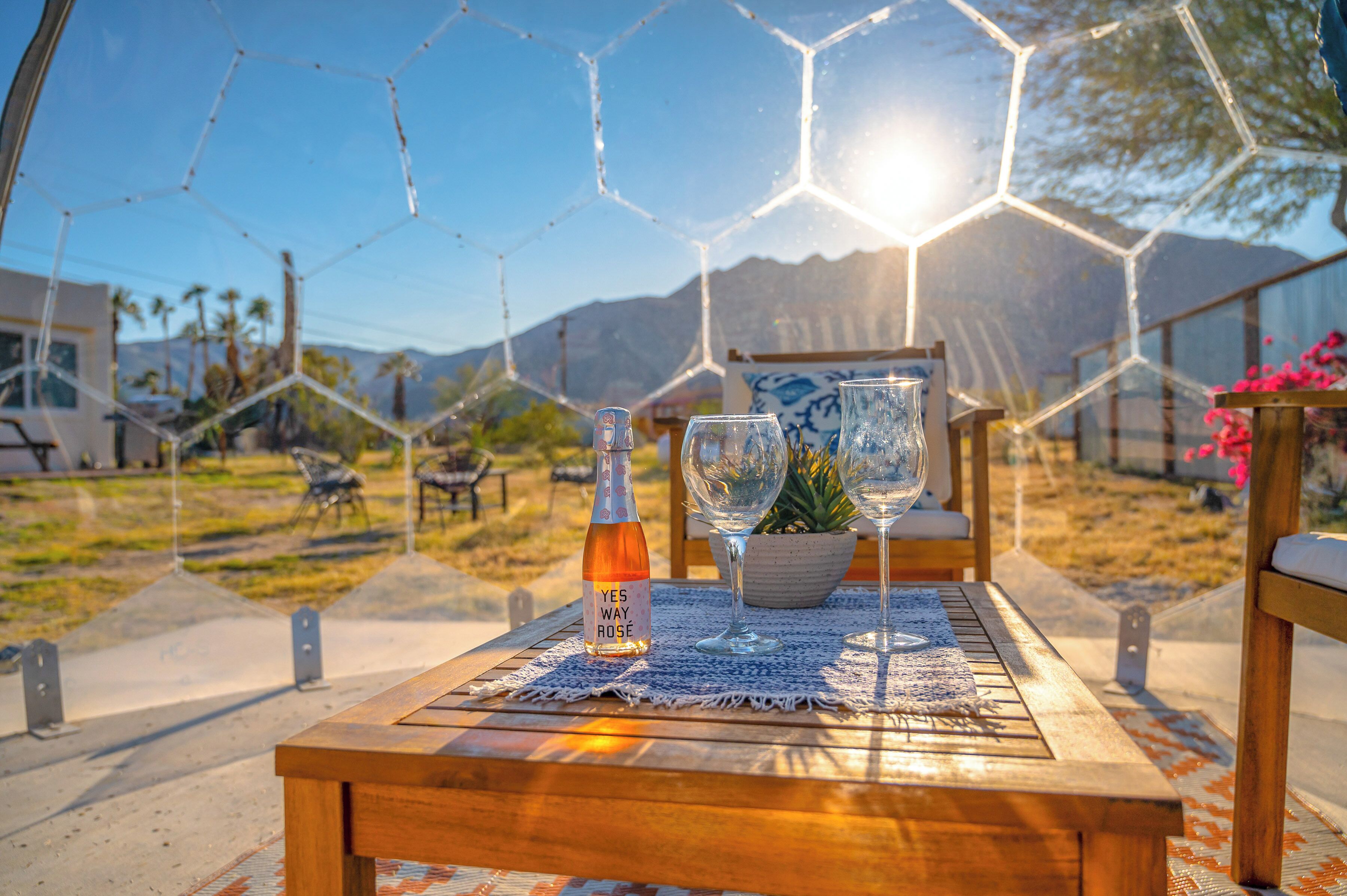 Una mesa de madera con una botella y vasos, colocada al aire libre bajo un cielo despejado y montañas al fondo.
