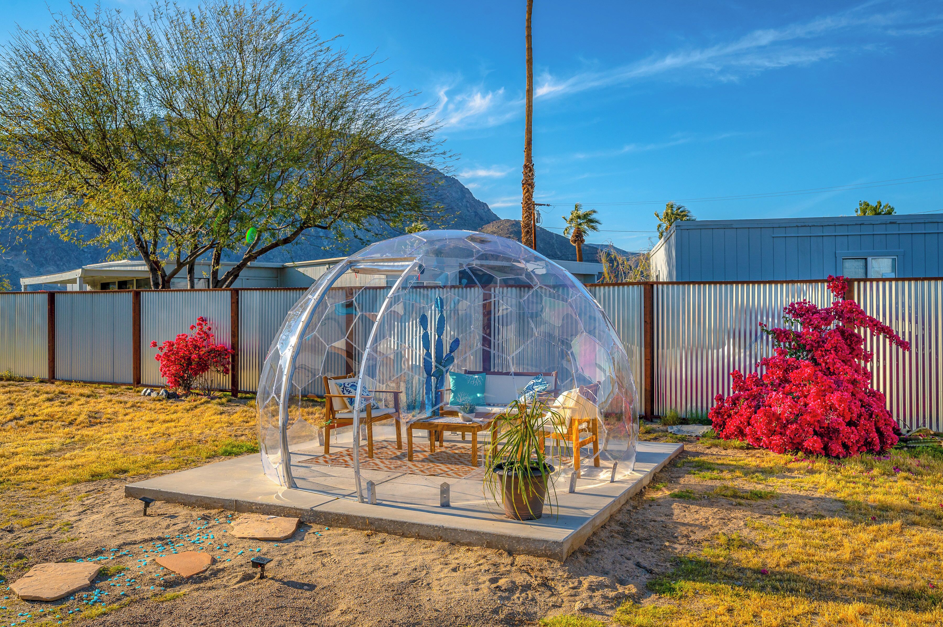 Estructura de cúpula transparente con asientos y una mesa, rodeada por un área cercada con vegetación desértica y una montaña al fondo.