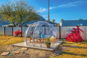 A transparent dome structure with seating and a table, surrounded by a fenced area with desert vegetation and a mountain in the background.