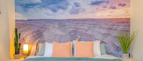 Un dormitorio con un mural grande de un cañón, una cama con varias almohadas, una lámpara de noche y una planta en maceta.
