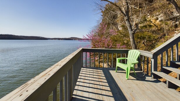 A wooden deck with a green chair, overlooking a calm lake with distant hills.