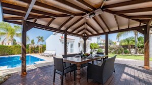 A covered outdoor area with a wooden deck, a table with chairs, and a view of a swimming pool and buildings in the background. - Thekla Oasis Villa - Steps from the Beach (Ayia Napa)