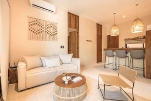 A modern living room with a sofa, a round coffee table, a woven bench, and a kitchen area with bar stools.