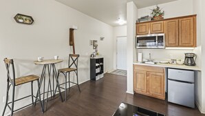 Una cocina con armarios de madera, un refrigerador y un microondas. Hay un pequeño comedor con dos taburetes y una mesa.