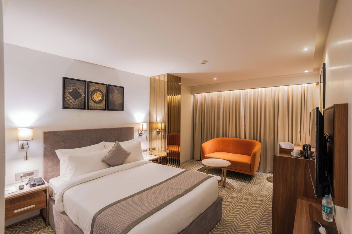 A hotel room with a large bed, a small orange armchair, a desk with a lamp, and a TV.