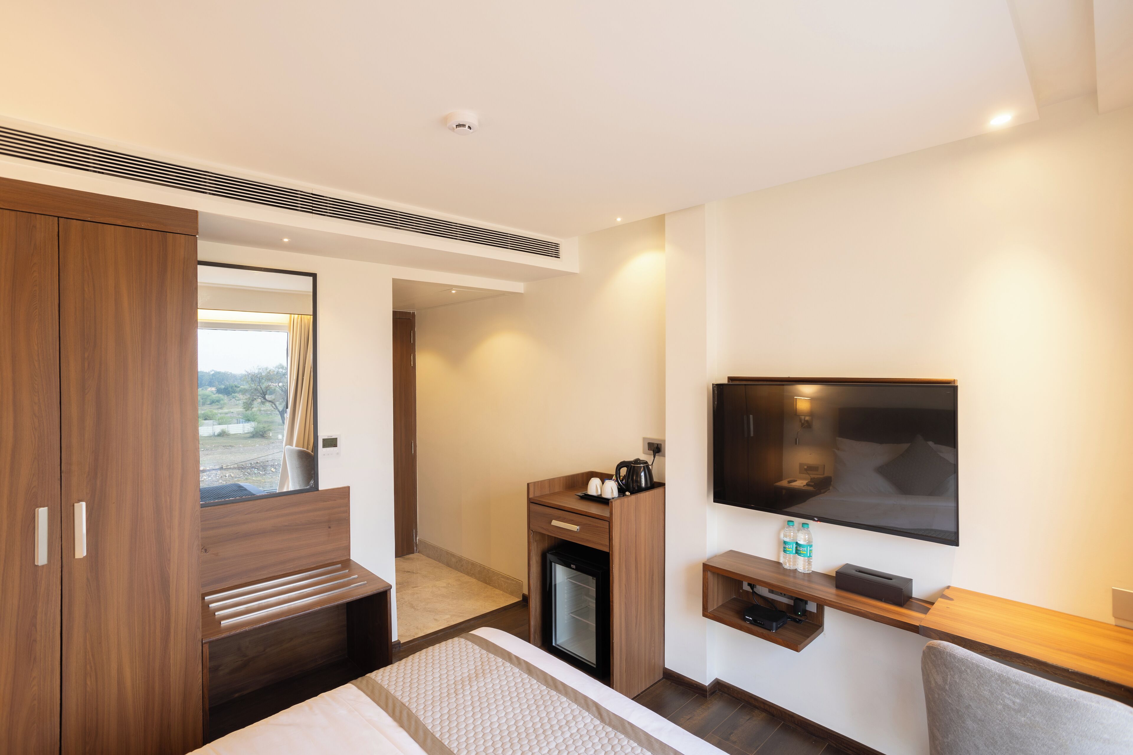 A modern hotel room with a bed, a TV, a mini-fridge, a desk, and a wardrobe.