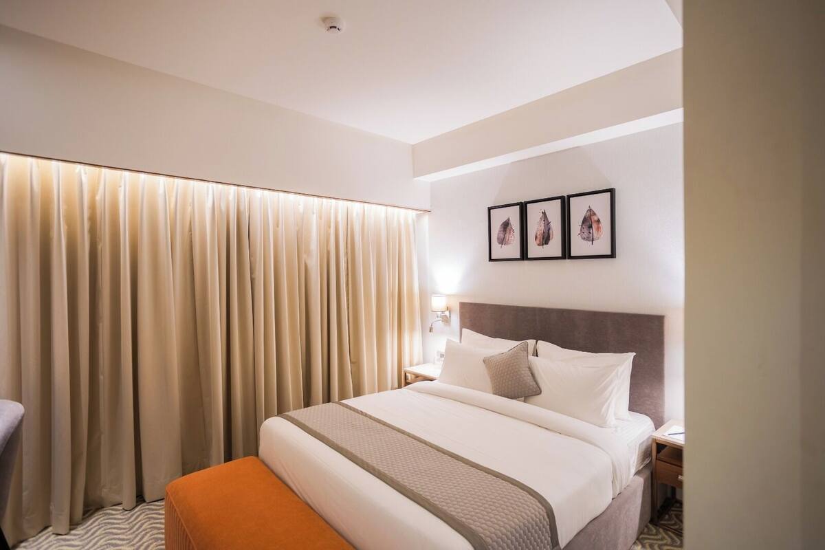 A hotel room with a large bed, a bedside table, a nightstand, a window with curtains, and three framed pictures on the wall.
