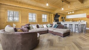 Een ruime woonkamer met een grote zithoekbank, een houten vloer met een herringbone patroon, en een keuken met een bar en barkrukken.