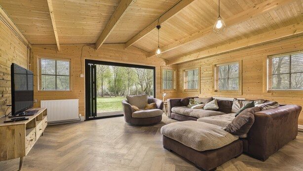 Een ruime woonkamer met houten wanden en vloeren, een grote modulaire bank, een ronde fauteuil en een televisie op een houten dressoir.