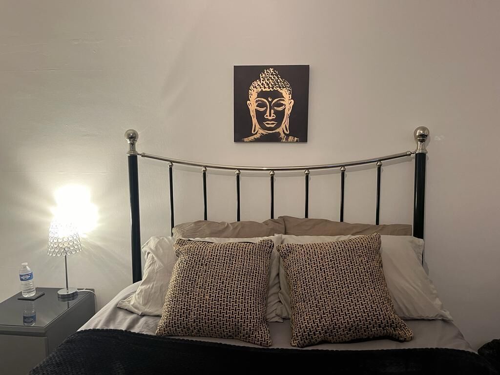 A bed with a metal headboard, two patterned pillows, a wall-mounted Buddha artwork, a bedside lamp, and a water bottle on a nightstand.