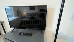 A flat-screen TV on a stand with a remote control, a bed, a window, and a wall-mounted mirror in the background. - Contractors - Families - Couples - 3x Bedrooms + Sofa Bed - Sleeps 8 M1 J26&25 (Ilkeston)