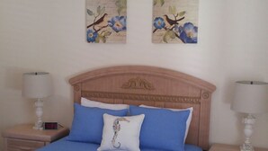 Een net opgemaakt bed met blauw bedlinnen, twee bijpassende kussens en een houten hoofdbord. Er staan twee nachtkastjes met lampen en een klein decoratief item op elk.