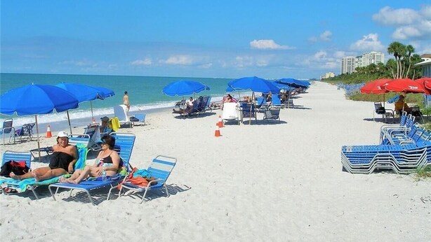 Een strand met rijen blauwe en rode ligbedden en parasols, een heldere lucht en een kalme zee.