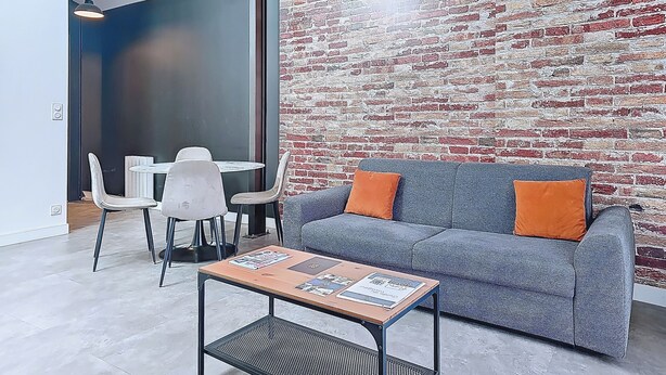 A modern living area with a grey sofa, orange cushions, a coffee table with magazines, a dining table with chairs, and a brick-patterned wall. - Appartement Le New-york, Chill & Relax ! (Cholet)