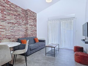 A modern living room with a brick wall, a grey sofa, a small table, a red ottoman, and a television. - Appartement Le New-york, Chill & Relax ! (Cholet)