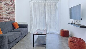 A modern living room with a brick wall, a grey sofa, a small table, a red ottoman, and a mounted TV.