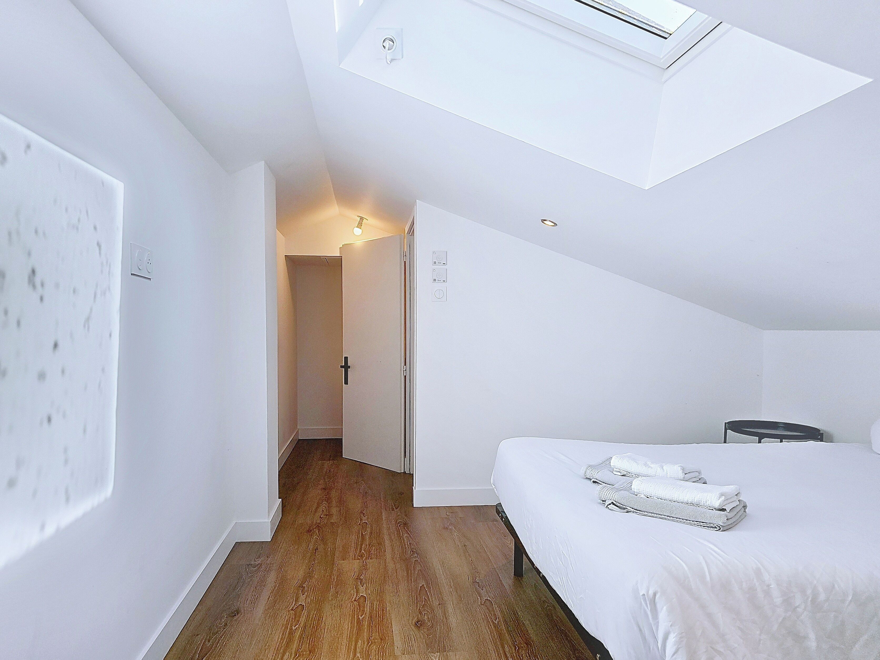 Ein Schlafzimmer mit Dachfenster, einem Bett, einem Nachttisch und einer Tür zu einem anderen Raum.