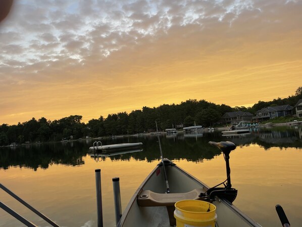 Un lago tranquilo al atardecer con barcos amarrados, un muelle en primer plano y casas al fondo.
