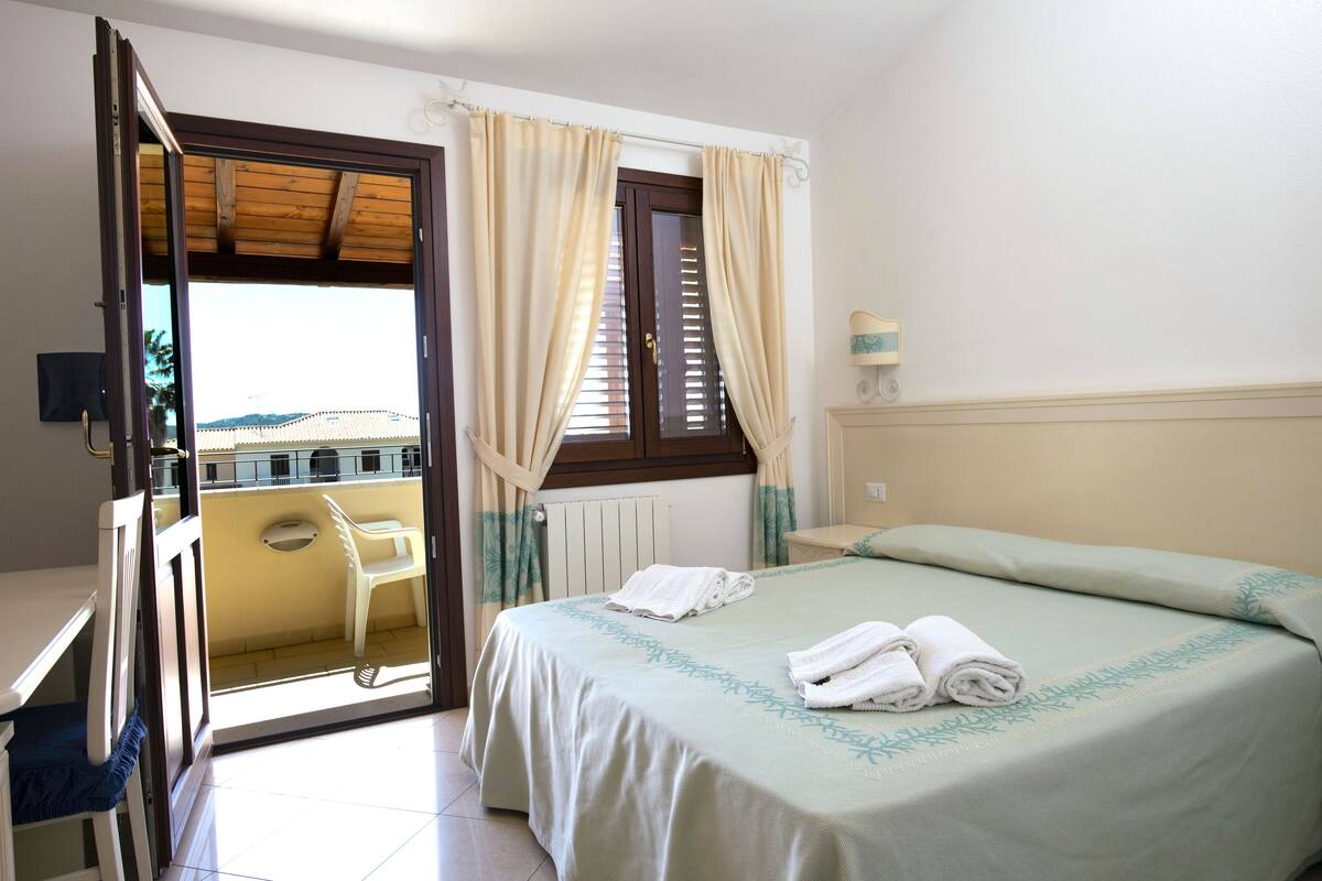 Um quarto com cama, janela com cortinas, radiador e vista para uma varanda.