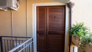 Una puerta de madera cerrada con el número 5, una barandilla metálica y una planta en maceta a su lado.