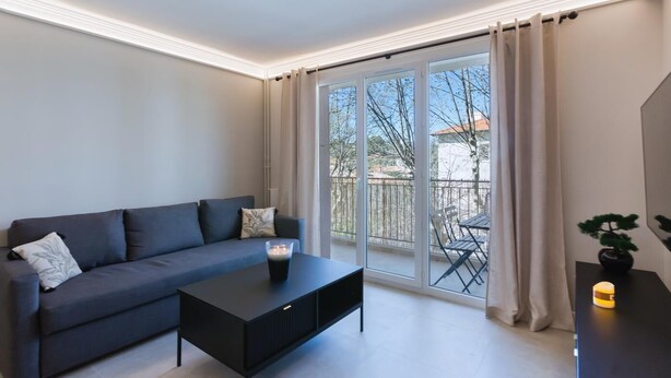 A modern living room with a dark grey sofa, a black coffee table, and a sliding glass door leading to an outdoor area with a table and chairs. - Antibes Calm: Your Coastal Haven (Antibes)