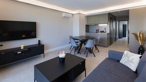 A modern living room with a flat-screen TV, a coffee table with a candle, a gray sofa, and a dining area with a round table and chairs. - Antibes Calm: Your Coastal Haven (Antibes)