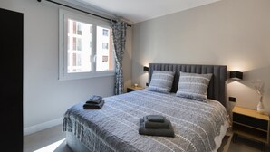 A modern bedroom with a large bed, a window with curtains, a bedside table with a lamp, and a nightstand. - Antibes Calm: Your Coastal Haven (Antibes)